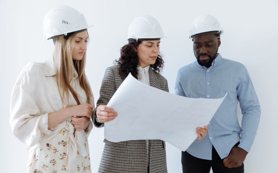 Three racially diverse real estate professionals — architect, engineer, and construction lead — reviewing blueprints together